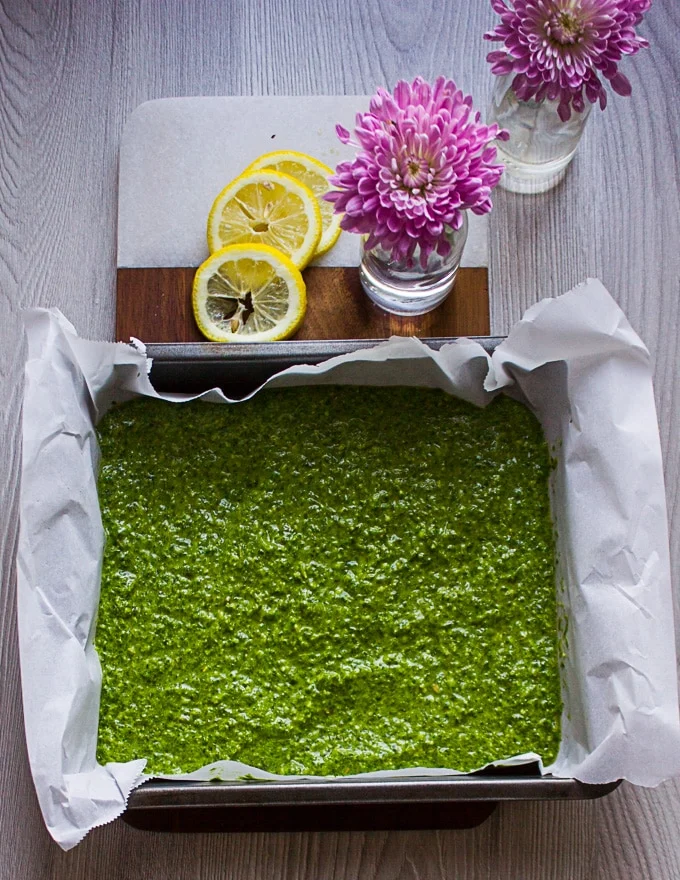 A batch of basil pesto spread over parchment paper in a baking pan and ready to go in the freezer surrounded by lemon slices