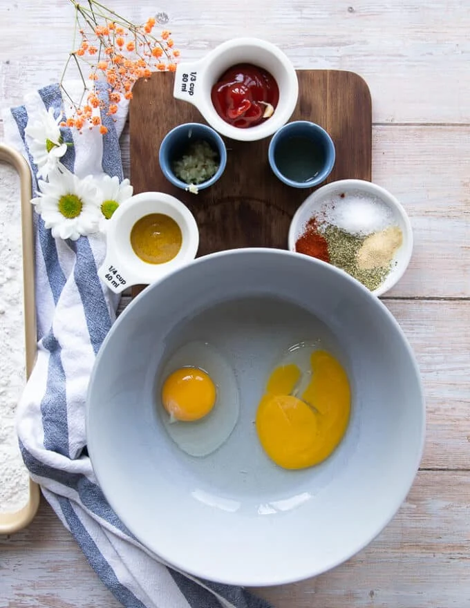 the wet coatings including a bowl of eggs, a bowl of mustard, a bowl of ketchup, minced garlic, lemon juice in a bowl, the same spice mix used for the dry coating,