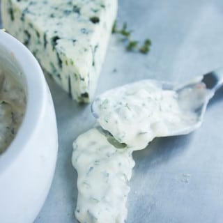 Blue cheese dip or bue cheese sauce spooned over a sheet by a spoon showing the consistency of it. A block of blue cheese in the background