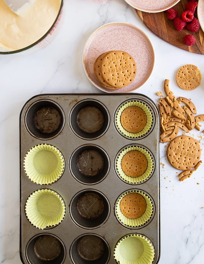 A muffin pan lined with baking sheets and a cookie pressed at the bottom of each paper to make the cheesecake base