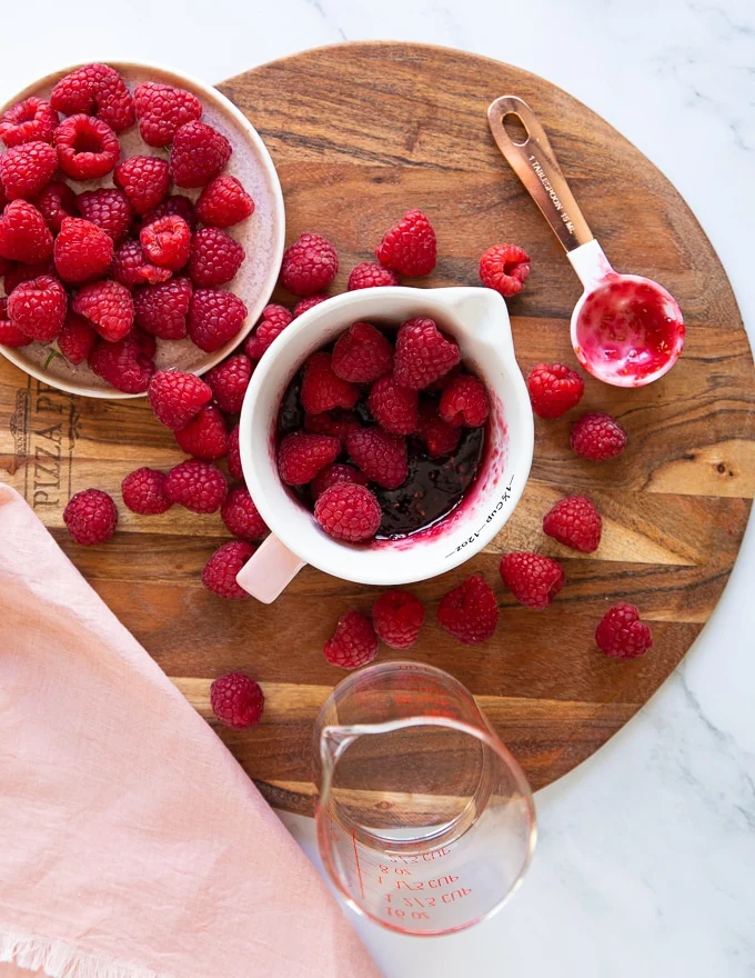 the cup of raspberry jam with hot water on the side and lots of fresh raspberries on a cutting board