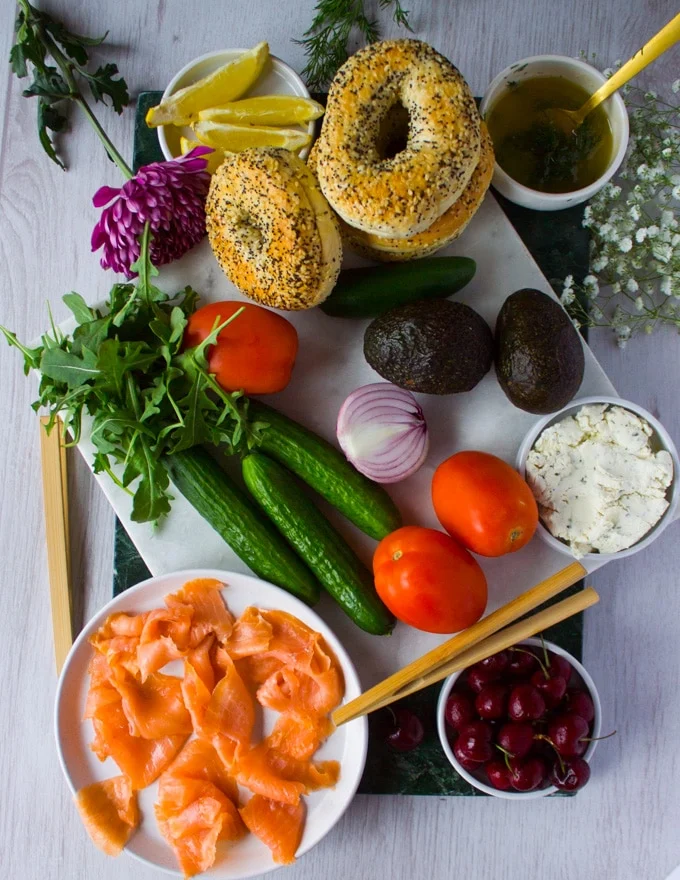 Whole Ingredients for smoked Salmon Platter on a white marble including smoked salmon, cucumber, avocados, bagels, dill, fruits, arugula