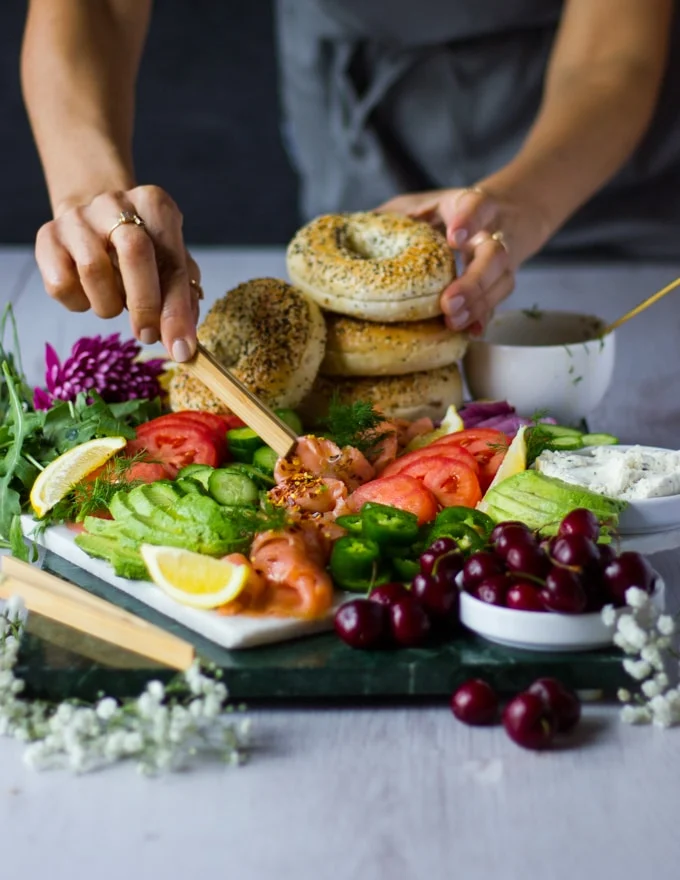 A hand holding the bagels while the other hands holding the smoked salmon using a tong to assemble the bagel and eat.