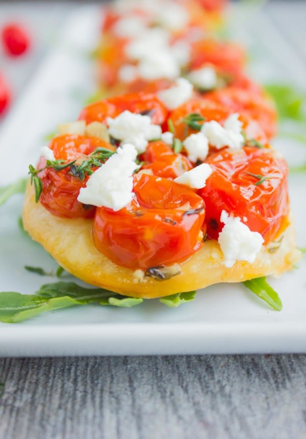 Mini Tomato Tartes Tatin sprinkled with crumbled feta cheese lined up on a while rectangular plate