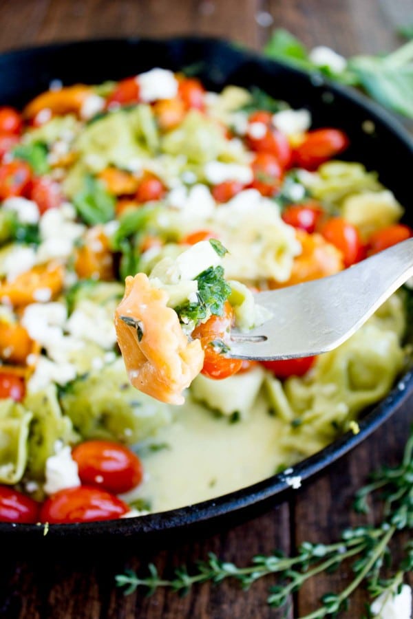 a fork with green and orange cheese tortellini in front of a skillet with cheese tortellini
