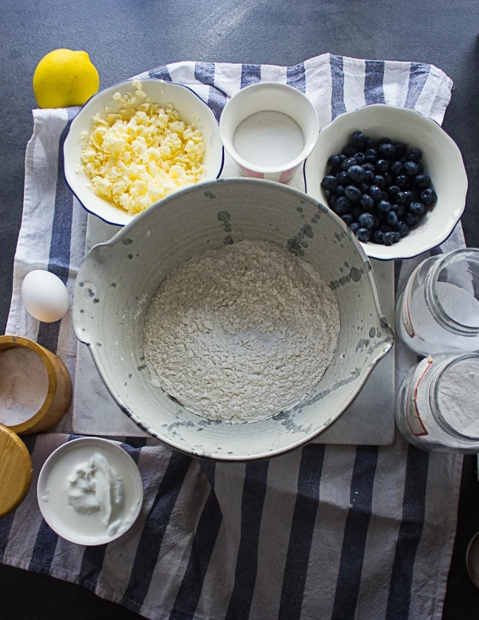 Ingredients for drop biscuits, a bowl of flour, some baking powder, an egg, grated butter, fresh blueberries, lemon, sour cream in a bowl