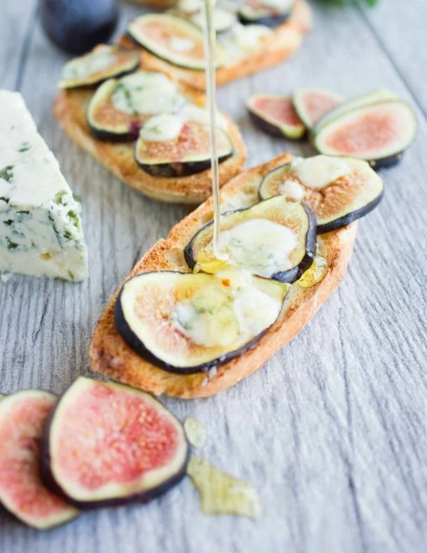 honey being drizzled on Fig Blue Cheese Honey Crostini placed on a rustic wood table with freshly sliced figs in the background