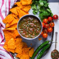 a metal bowl of salsa surrounded by chips, fresh tomatoes, jalapeno and cilantro over a tray