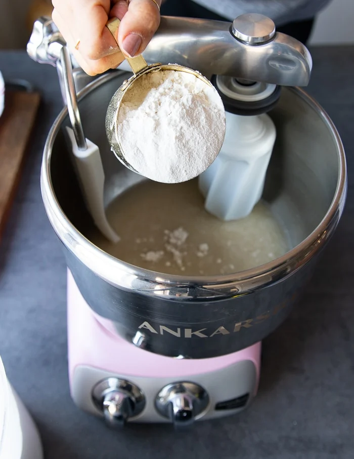 A hand pouring in the flour gar4dually into a stand mixer to make the pizza dough recipe