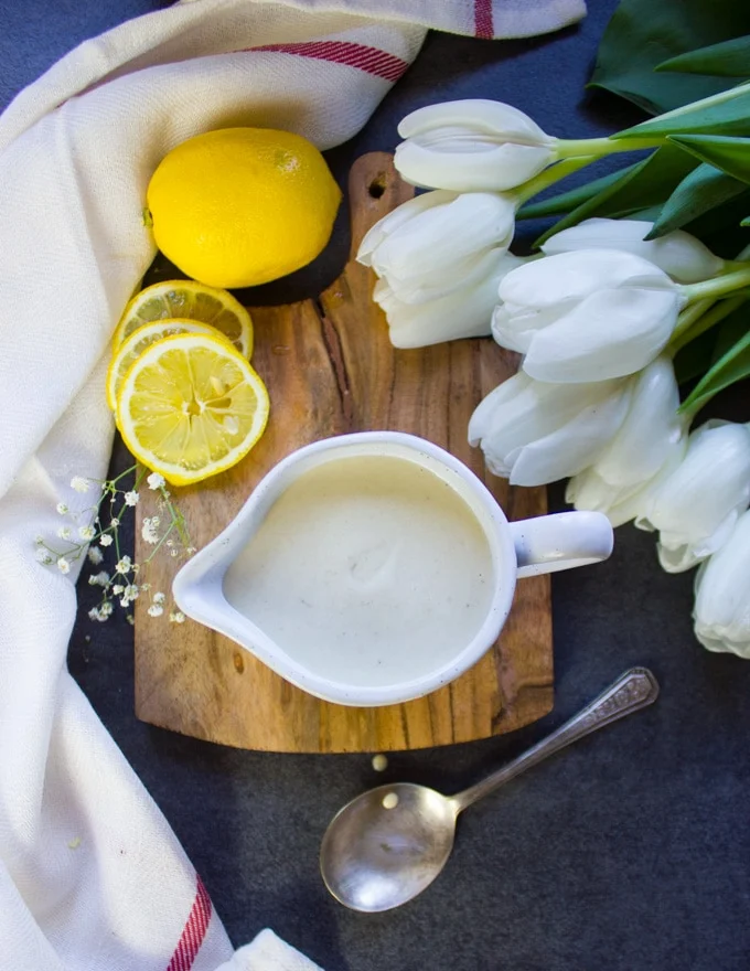 The tahini sauce in a pouring cu with lemon slices around it and white flowers, and a tea towel