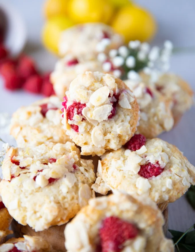 stacked up raspberry muffins over one another with fresh rasberries around them and some lemons in the background on a white board