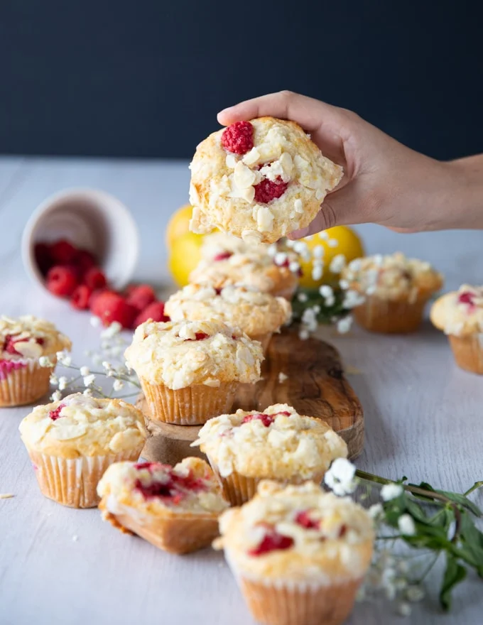 A hand holding a raspberry muffin above a pile of muffins to show the raspberries in tact and the crunch topping