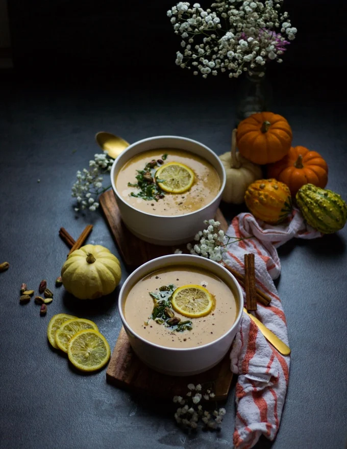two bowls of mediterranean soup of autumn squash with a tea towel, surrounded by squash and lemon slices and cinnamon sticks