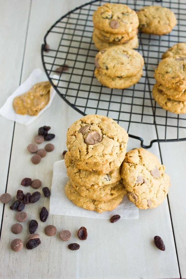 Peanut Butter Chocolate Chip Cookies stacked on top of each other on a piece of parchment paper with some chocolate chips on the side and more cookies on a cooling rack in the background