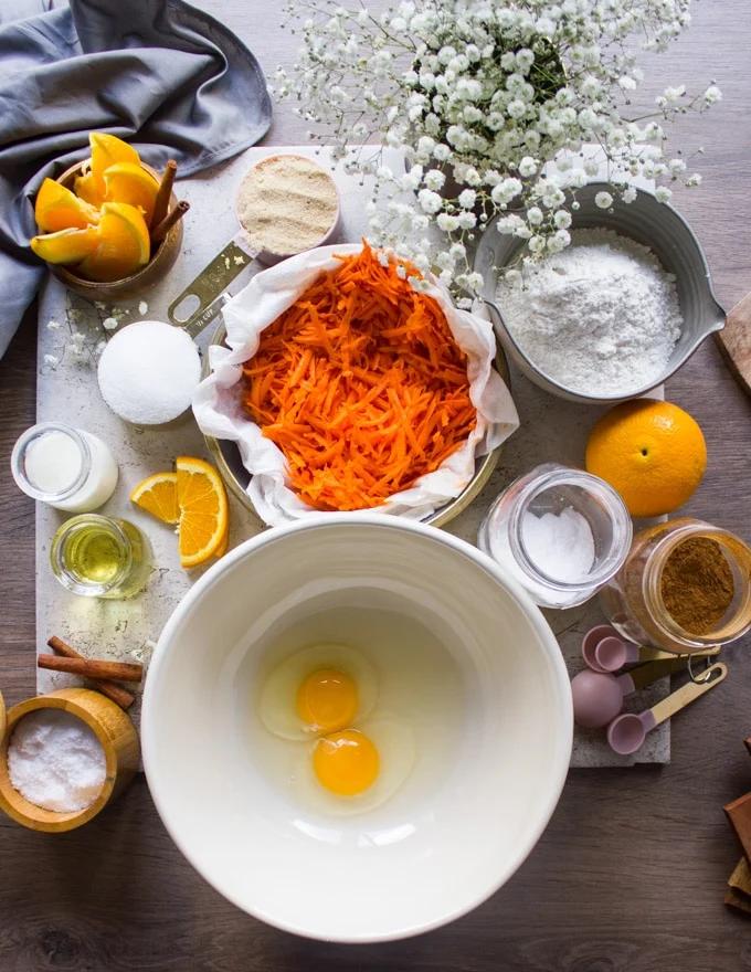 different bowls with ingredients for the carrot cake, a bowl of eggs, a bowl of carrots, oil, a bowl of sugar, a bowl of flour, baking soda, salt, cinnamon, buttermilk, orange juice and some fresh oranges