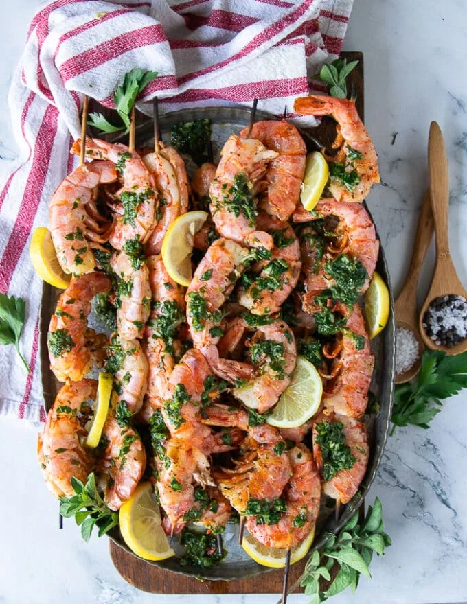 Final plate of grilled shrimp with a tea towel on a cutting board surrounded by lemon slices, fresh herbs and some chilli flakes
