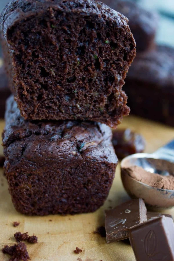 2 stacked loafs of Double Chocolate Zucchini Bread on a wooden board with some chocolate and cocoa powder on the side