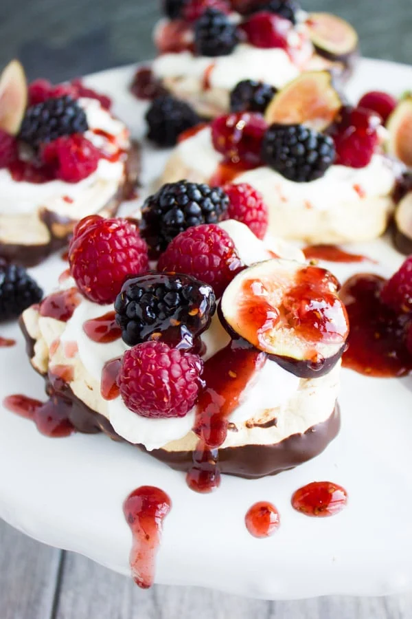 Chocolate Dipped Mini Pavlova drizzled with raspberry sauce and decorated with fresh berries and fig slices on a white cake stand
