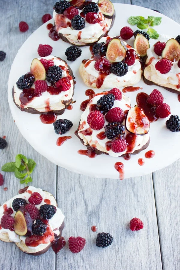Chocolate Dipped Mini Pavlova drizzled with raspberry sauce and decorated with fresh berries and fig slices on a white cake stand
