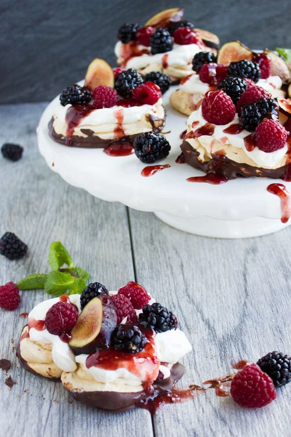 Side view of Chocolate Dipped Mini Pavlova drizzled with raspberry sauce and decorated with fresh berries and fig slices on a white cake stand