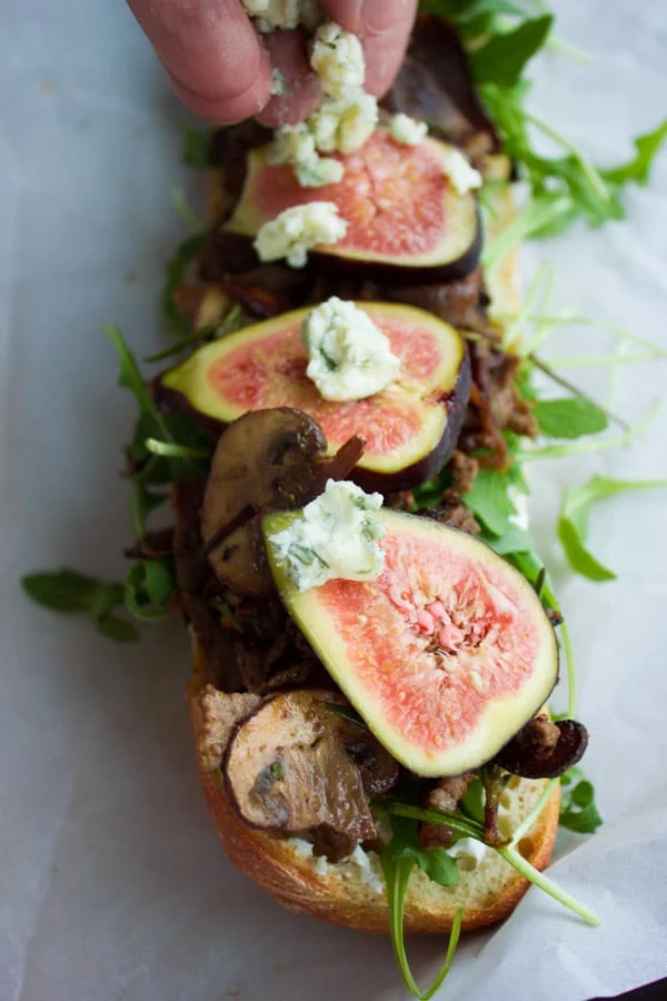blue cheese being sprinkled on top of an open baguette sandwich with steak slices, arugula and fresh figs