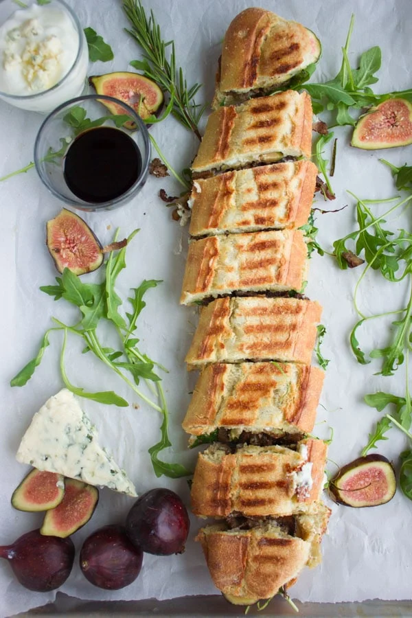 overhead-shot of a filled sandwich baguette cut into slices and whimsically arranged on parchment paper with arugula, figs and a wedge of cheese