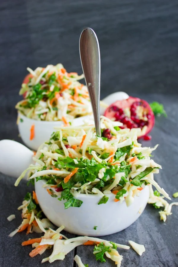 Side view of Coleslaw with Kale, Apples and Pomegranate with a fork sticking out of the front dish and some slaw falling out of the bowl onto the table.