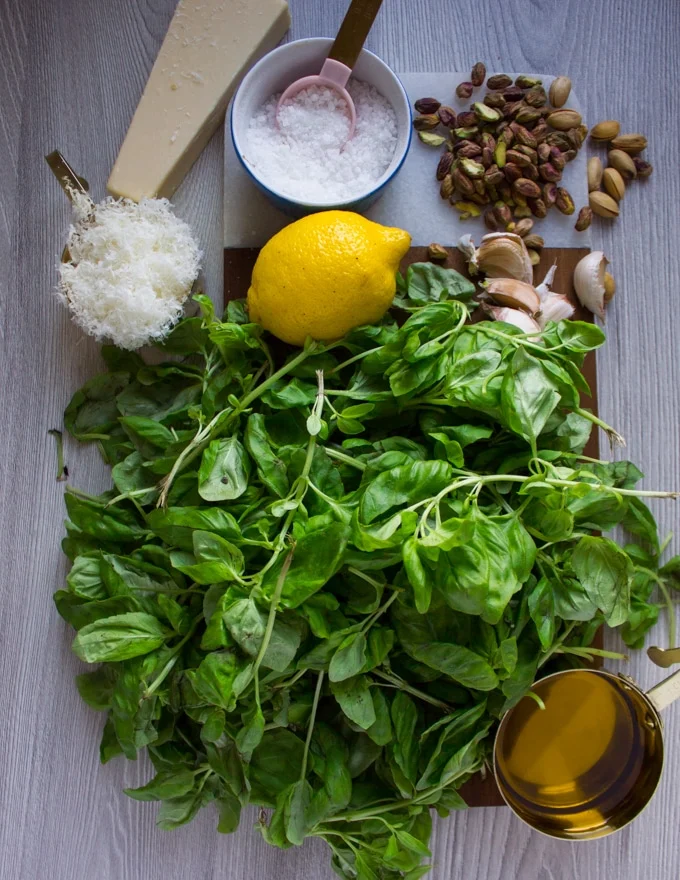 Ingredients on a cutting board: parmesan cheese, salt, pistachios, lemon, basil, spinach, olive oil.
