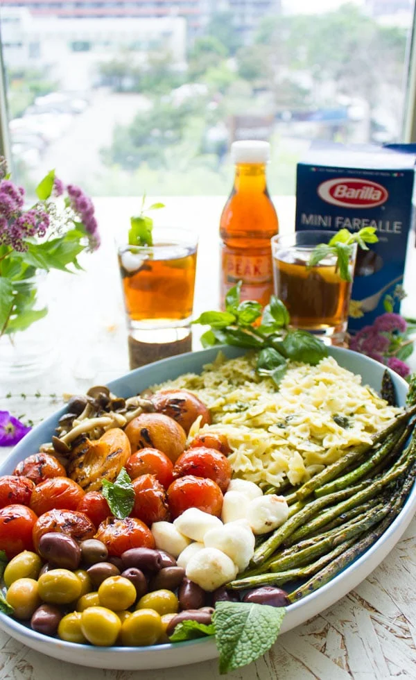 Side view of summer pasta salad plated in an oval dish with grilled vegetables, asparagus and mini mozzarella. Glasses of sweet tea and a box of farfalle pasta in the background.