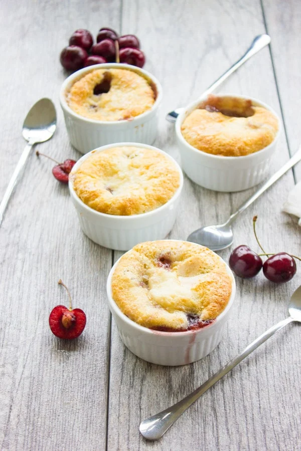 close-up of four small ramekins with Sweet Cherry Buckle next to some fresh cherries and dessert spoons