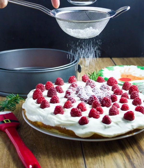 powdered sugar being sifted on top of a Fluffy White Raspberry Holiday Cake. Light as air fluffy tender and moist white cake with vanilla cream and fresh raspberries.