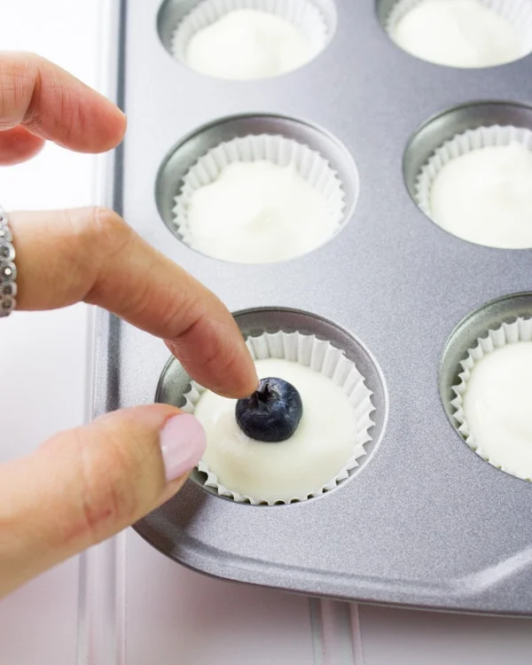 a blueberry being pushed into Greek yogurt cheesecake filling in a mini muffin tin