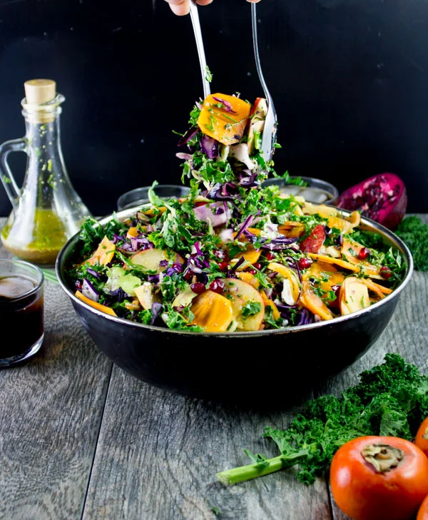 Kale Salad with apples, persimmon and pomegranate seeds being tossed in a big black salad bowl