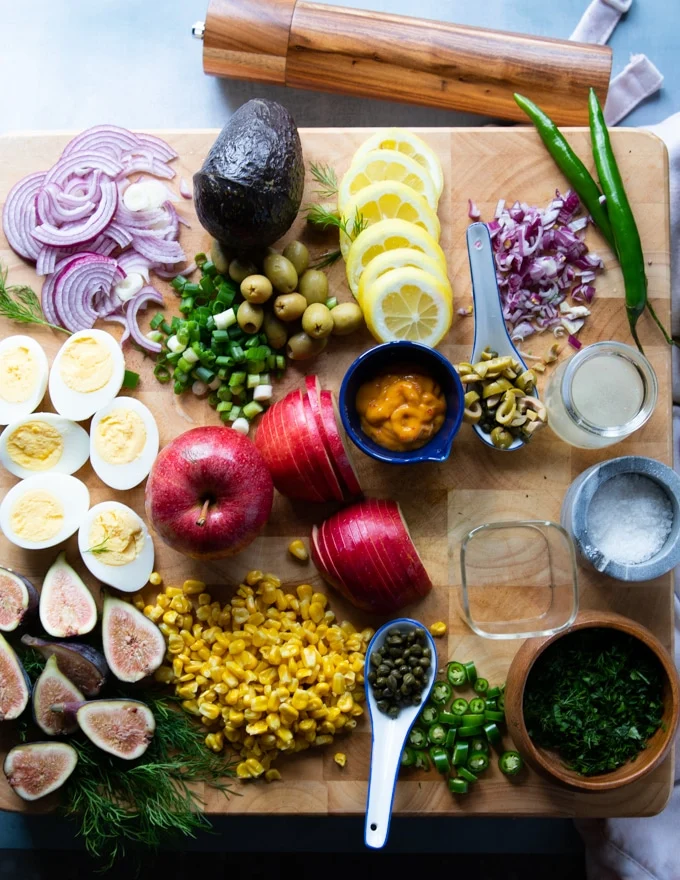 Ingredients on a wooden board for the tuna salad recipe including boiled eggs, apples, avocado, lemon juie, onions, olives, olive oil, capers, fres herbs, tomatoes, corn, tuna, jalapenos