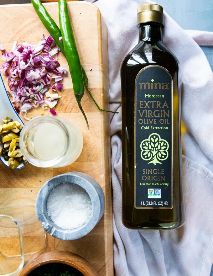 a bottle of olive oil on a board next to the olive oil salad ingredients such as lemon juice, salt, onions, minced olives