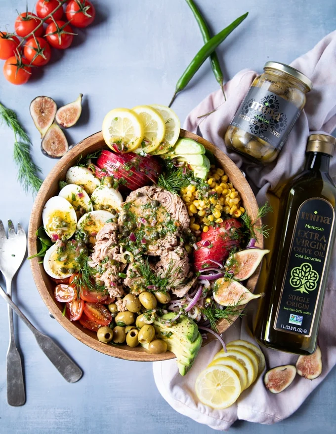 finished tuna salad recipe with the bottle of olive oil and green olives on the side. Some fresh figs on the table, a serving spoon and fresh dill. The tuna salad recipe is loaded with tuna, hard boiled eggs, avocados, tomatoes, apples, corn and tomatoes