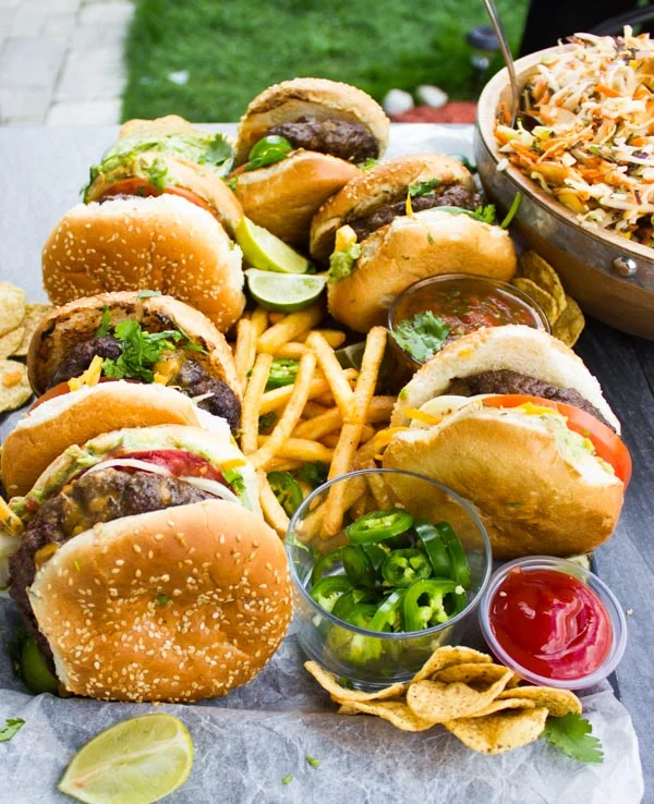 Side view of Lamb Burgers Loaded with Nacho toppings on a plate with french fries, jalapeno slices and guacamole.