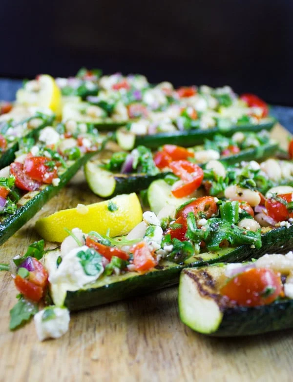 Side view of grilled Zucchini Boats loaded with White Bean Salad served on a wooden chopping board with some lemon slices on the side.