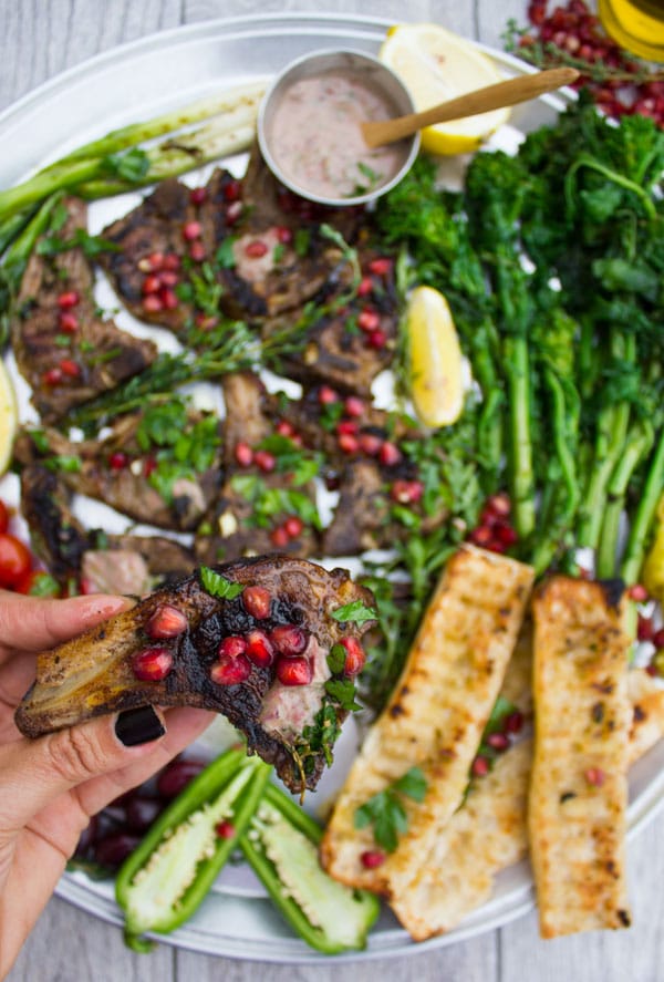 a hand holding a succulent lamb chop sprinkled with pomegranate seeds with a platter with lamb chops and grilled veggies in the background