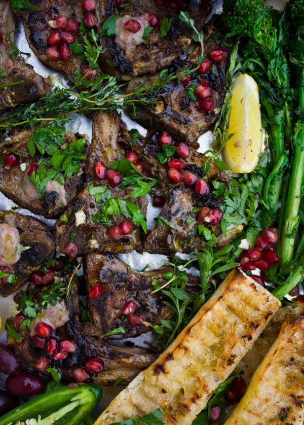 Succulent Grilled Lamb Chops with Black Olive Herb Butter sprinkled with pomegranate seeds and arranged on a silver platter with grilled rapini as well as some charred garlic baguette.