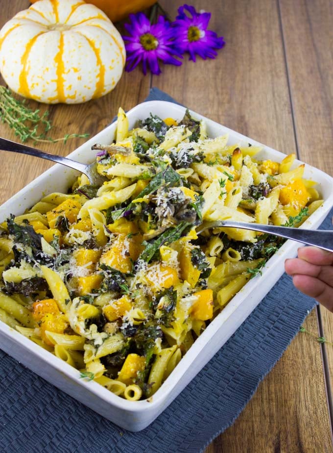 Easy Baked Pasta with Roasted Pumpkin, Kale and Mushrooms in a casserole on a blue place mat with a pumpkin in the background