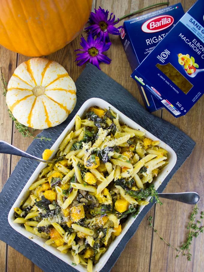 Easy Baked Pasta with Roasted Pumpkin, Kale and Mushrooms in a casserole on a blue place mat with a pumpkin and more pasta in the background