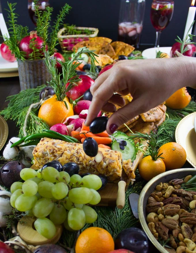 a hand holding an olive with a huge display of cheeses and fruit in the background