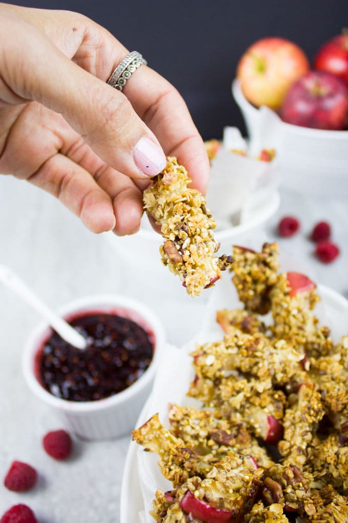 a hand holding a baked apple fry over a dish with more apple fries and fresh raspberry sauce