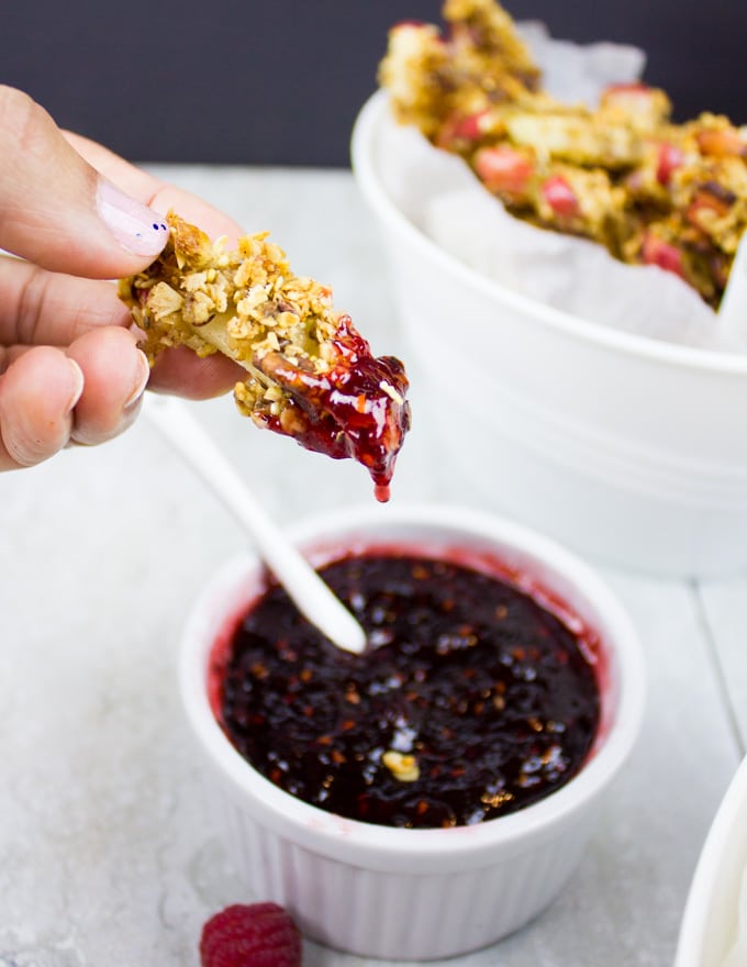 granola-coated baked apple fry just being dipped into a small white bowl with raspberry sauce
