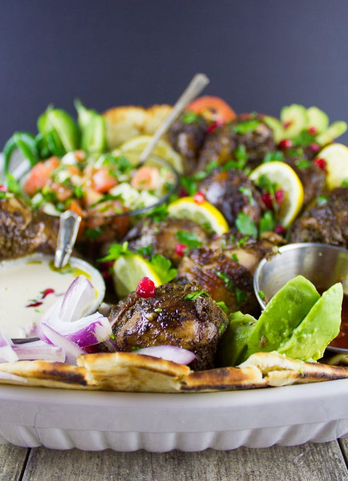 side view of Chicken Shawarma Platter served on charred naan bread with Tahini Sauce, Harissa Sauce and Tomato Feta salad in small dishes nestled in the chicken