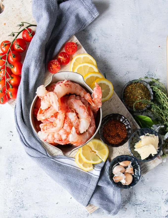 Ingredients to make shrimp pasta including raw shrimp, a bowl of seasoning, some lemon slices, garlic slices, fresh herbs, baby tomatoes