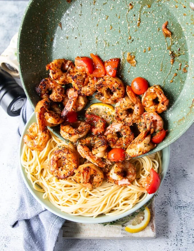 The cooked shrimp and sauce is being poured over a plate of cooked pasta to toss around