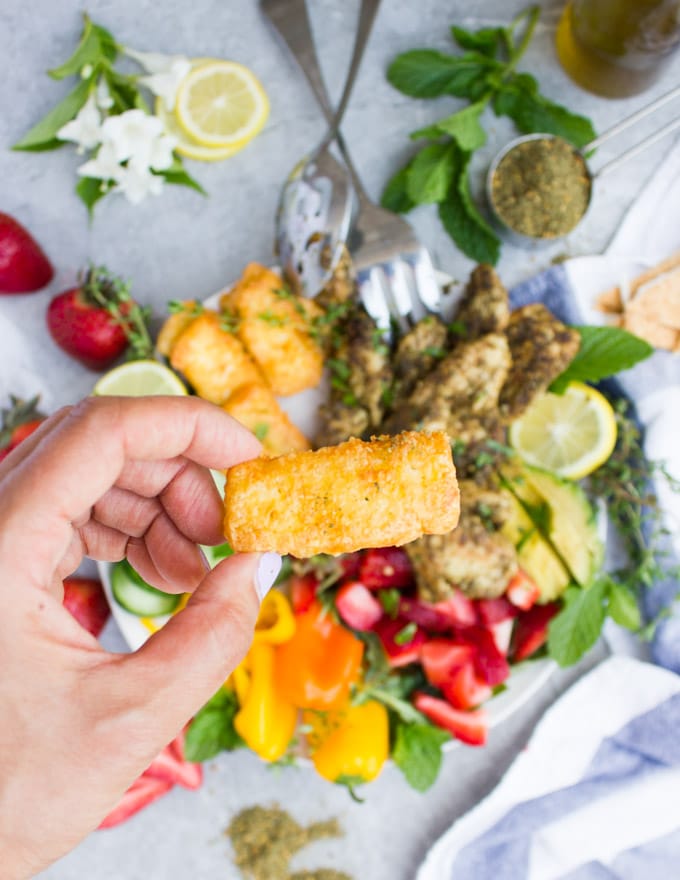 a hand holding a fried halloumi cheese stick over a bowl of za'atar chicken salad