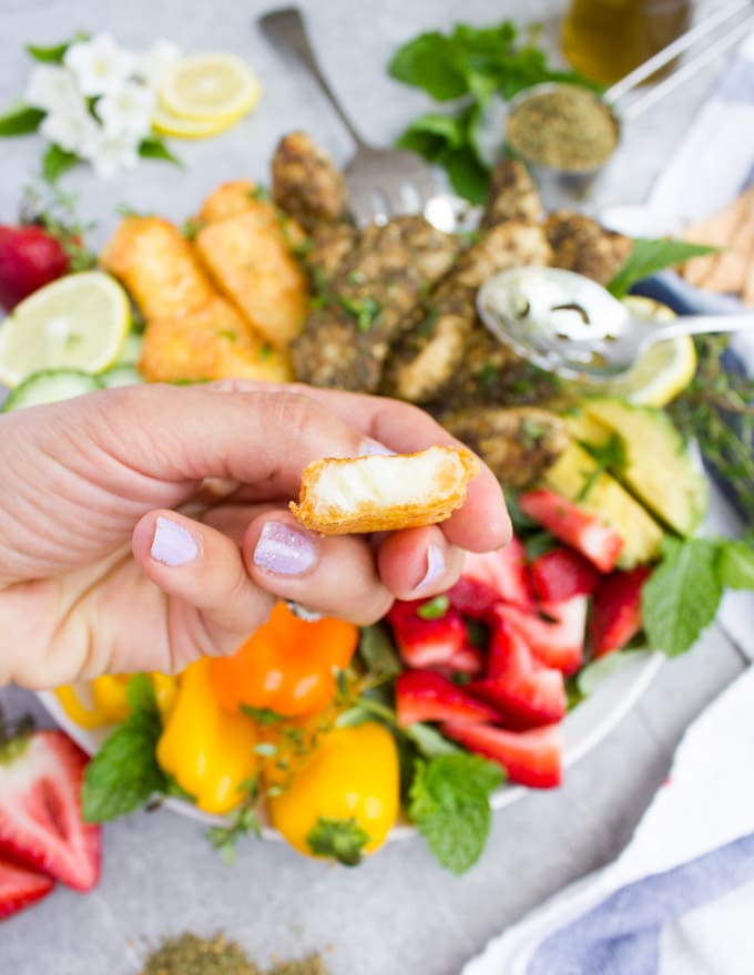 a hand holding half a piece of fried halloumi cheese over a bowl of chicken and halloumi salad